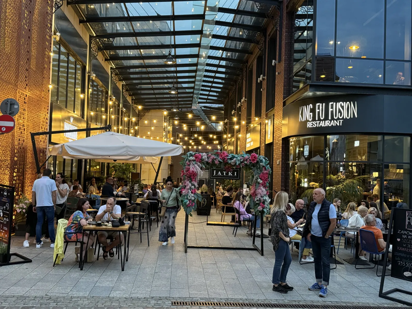 Wejście do pasażu Jatki w Bydgoszczy o zmierzchu – szklany dach, girlandy świateł, kwiatowa brama Jatki i narożnik restauracji King Fu Fusion z ogródkiem