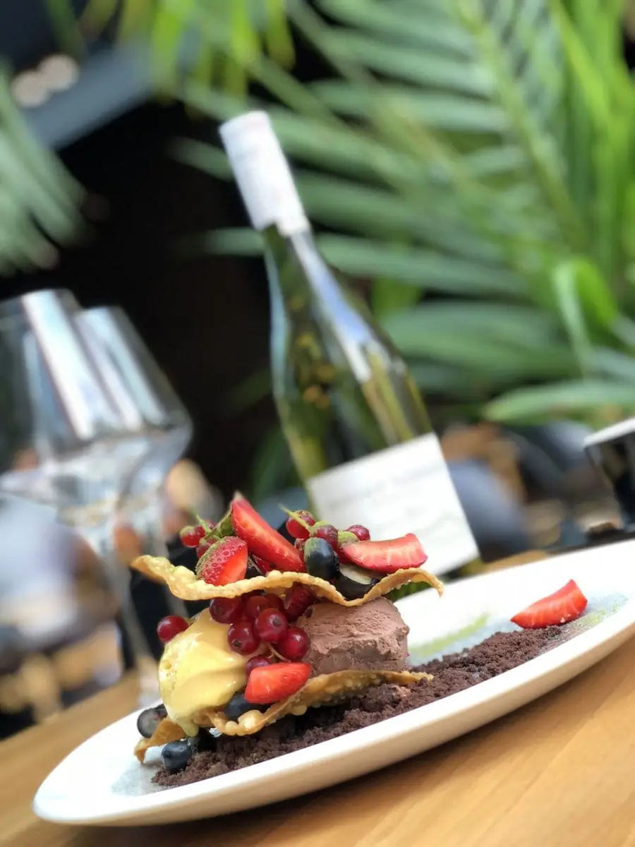 Deser z lodami czekoladowymi i mango, truskawkami, czerwoną porzeczką i borówkami między chrupiącymi płatkami, z butelką białego wina w tle – King Fu Fusion Bydgoszcz