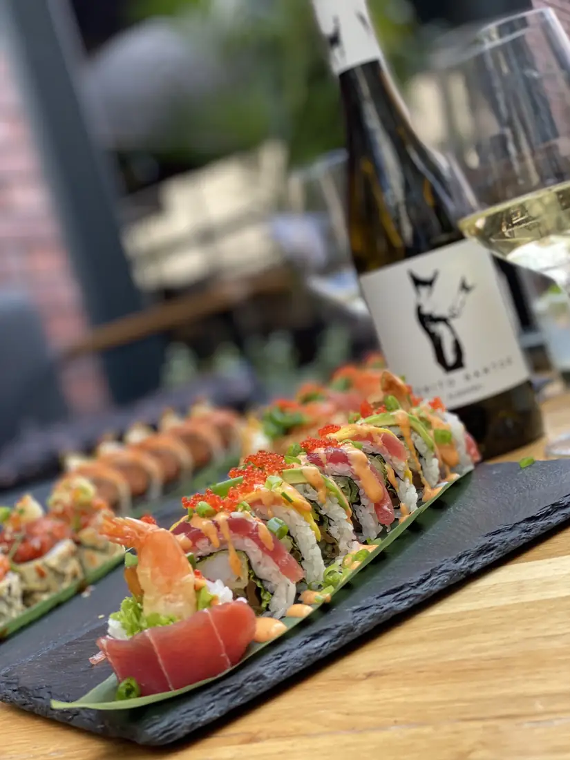 Rolki sushi fusion z tuńczykiem, krewetką w tempurze, awokado i kawiorem tobiko na łupku, w tle butelka białego wina – King Fu Fusion Bydgoszcz