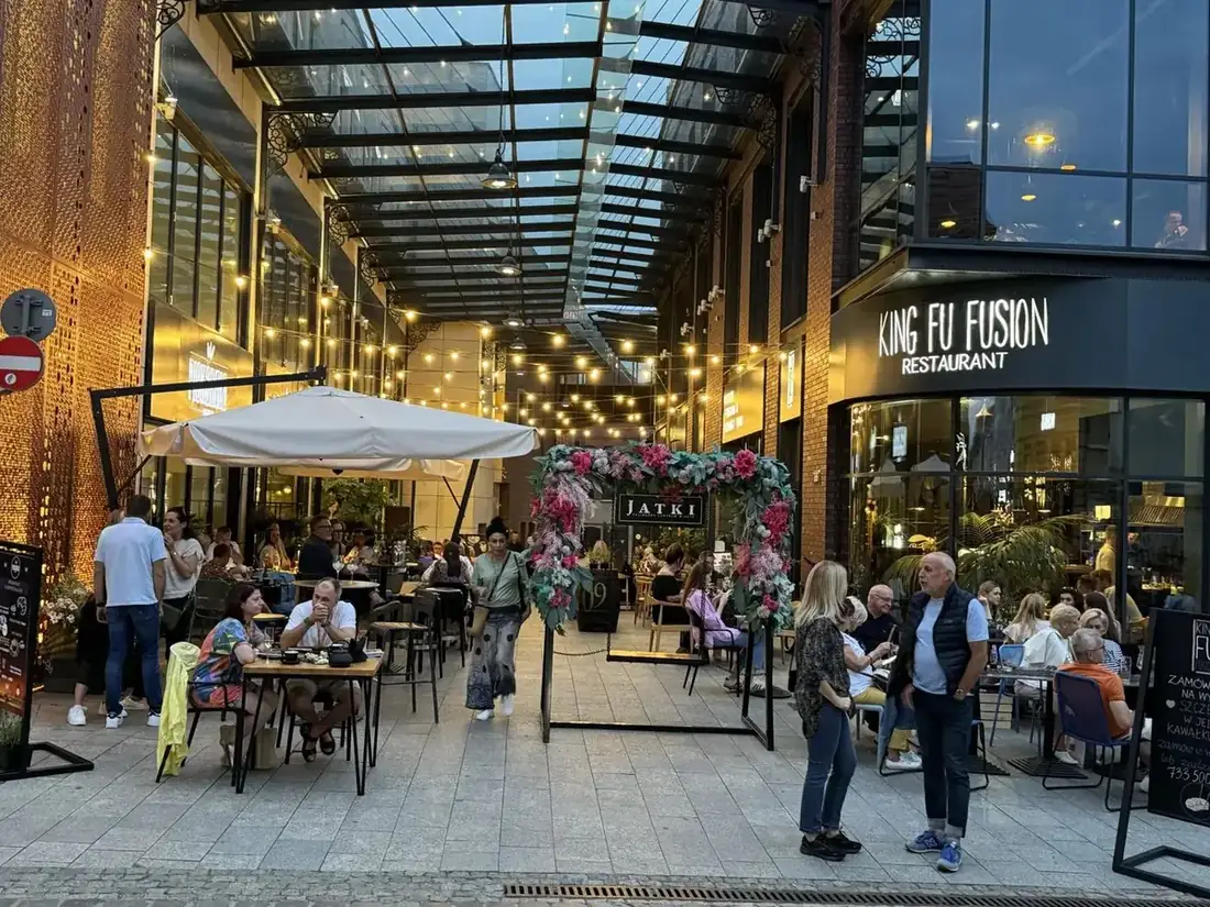 Wejście do pasażu Jatki w Bydgoszczy o zmierzchu – szklany dach, girlandy świateł, kwiatowa brama Jatki i narożnik restauracji King Fu Fusion z ogródkiem